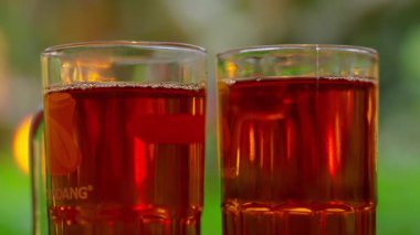 Koyu kırmızı içecekle dolu iki bardak, muhtemelen çay, görünür köpüklü. Arka planda bulanıklık var, yeşillik ve açık hava manzarası var..