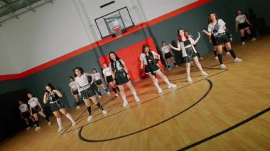 Bir spor salonunda gösteri yapan bir grup genç dansçı. Koordineli kıyafetler giyiyorlar, koreografili bir gösteri sergiliyorlar. Arka plandaki basketbol potası spor temasına ekleniyor.