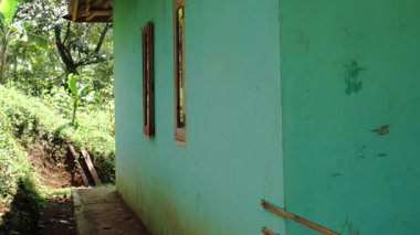 Bereketli yeşilliklerle çevrili, ahşap pencereli turkuaz renkli bir evin yan görüntüsü. Zemin biraz çamurlu, bu da kırsal bir alanı gösteriyor..