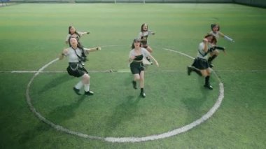 Altı dansçıdan oluşan bir grup futbol sahasında senkronize bir gösteri yapıyor. Sportif kıyafetler giyiyorlar, kolları havada enerjik pozlar veriyorlar. Yeşil çim ve dairesel alan işaretleri performansları için canlı bir zemin oluşturuyor..