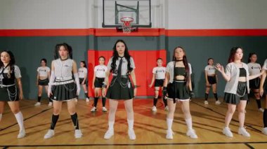 Bir grup dansçı bir spor salonunda koreografi gösterisi yapıyor. Şık kıyafetler giyiyorlar, etek ve spor kıyafetleri karışımı, enerjik hareketler sergiliyorlar. Basketbol potası arka planda görünür, atletizme katkı sağlar.