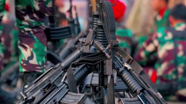 Arka planda kamuflaj üniformalı askerlerle birlikte askeri tüfeklerin yakın plan görüntüsü. Sahne bir hazırlık ve askeri varlık hissi veriyor..