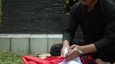 Siyah elbiseli bir kişi kırmızı ve beyaz bir bayrak tutuyor, muhtemelen bir tören ya da kutlamaya hazırlanıyor. Arkaplanda yeşillik ve desenli bir duvar var..