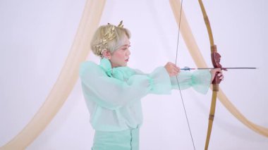 Nane yeşili bir kıyafet giymiş, okla yay doğrultmuş biri. Dekor, arka planda yumuşak perdeler içererek sakin bir atmosfer yaratıyor. Bireyin kısa sarı saçları ve dekoratif başlığı var..