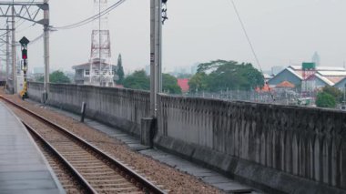 Beton duvar ve yeşilliklerle çevrili tren rayları olan bir demiryolu platformu. Sahne bulutlu, arka planda binalar ve bir telekomünikasyon kulesi var..