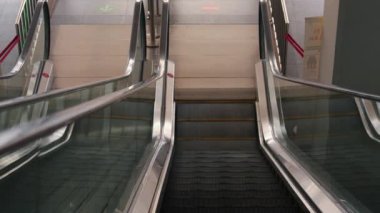 İki yürüyen merdivenin görüntüsü, biri yukarı çıkıyor, diğeri aşağı iniyor, modern bir iç mekan ortamında. Yürüyen merdivenler metal ve camdan yapılmış, temiz ve parlak bir görünüme sahip. Zemin kiremitlerle kaplı ve yön işaretleri görülebiliyor..