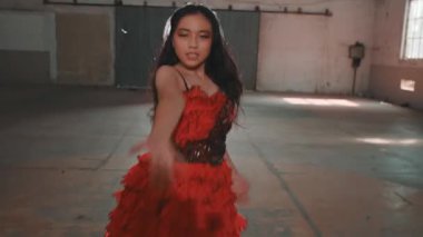 Parlak elbiseli genç bir kadın endüstriyel alanda dans ediyor. Saçları zarifçe hareket ederken, neşeli ifadesini ve canlı kıyafetini sergiliyor..