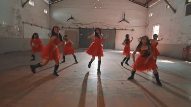 Kırmızı elbiseli bir grup dansçı boş bir depoda koreografi gösterisi yapıyorlar. Dansçılar çeşitli pozlarda, hareket ve enerji gösteriyorlar..