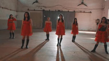 Kapalı bir mekanda altı dansçıdan oluşan bir grup sahne alacak. Canlı kırmızı elbiseler ve siyah çizmeler giyiyorlar, enerjik dans figürleri sergiliyorlar. Arka planda endüstriyel elementler var, dinamik atmosferi geliştiriyor..