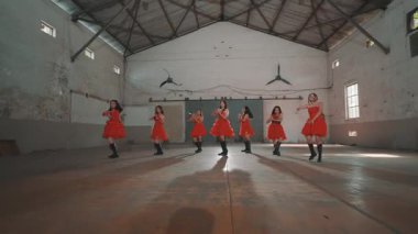 Bir grup kırmızı elbiseli dansçı endüstriyel alanda gösteri yapıyorlar. Dansçılar senkronize, onları aydınlatan spot ışıklarıyla dinamik hareketler sergiliyorlar. Arka planda, yüksek tavanlı ve vantilatörlü boş bir köy salonu yer alıyor..