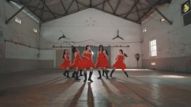 Sanayi bölgesinde kırmızı elbiseli beş dansçıdan oluşan bir grup. Dansçılar dinamik bir pozda, koreografilerini sergiliyorlar. Arka planda yüksek tavanlı ve asgari dekorlu geniş bir açık alan yer alıyor..