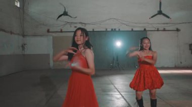Kırmızı elbiseli iki genç kadın endüstriyel alanda dans ediyor loş ışık ve spot ışıklarıyla.