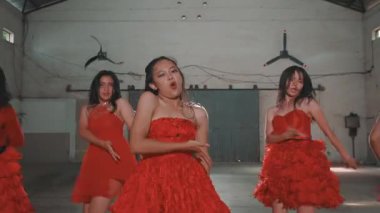 Canlı kırmızı elbiseler giyen bir grup genç kadın, geniş bir iç mekanda enerjik bir şekilde dans ediyorlar. İfadeleri birlikte performans sergilerken neşe ve heyecan gösteriyor..