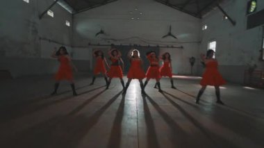 Kırmızı elbiseli altı dansçı yüksek tavanlı ve geniş pencereli endüstriyel bir alanda gösteri yapıyor. Gölgeleri yere doğru uzanıyor ve dramatik bir etki yaratıyor..