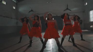 Bir grup genç dansçı loş bir odada canlı kırmızı elbiseler giyerek gösteri yapıyorlar. Enerjik atmosfer dinamik pozları ve hareketleriyle vurgulanır. Canlı bir dans performansı sergilerler..