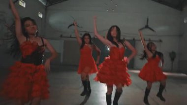 Bir grup kırmızı elbiseli dansçı endüstriyel alanda gösteri yapıyorlar. Baş dansçı kendinden emin bir şekilde önde duruyor, arkasında başkaları var, hepsi de dinamik pozlar veriyor..