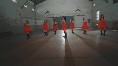 Bir grup kırmızı elbiseli dansçı yüksek tavanlı ve ahşap zeminli endüstriyel alanda gösteri yapıyorlar. Atmosfer enerjik ve dinamik, senkronize hareketlerini gösteriyor..