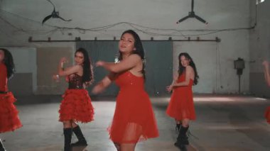 Dört genç bayan dansçıdan oluşan bir grup geniş, endüstriyel tarzda bir mekanda gösteri yapıyorlar. Canlı kırmızı elbiseler giyiyorlar, senkronize dans figürleri sergiliyorlar. Arka planda büyük hayranlar ve dokulu bir duvar var.