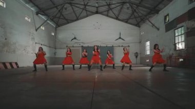 Bir grup kırmızı elbiseli dansçı yüksek tavanlı ve geniş pencereli endüstriyel bir alanda gösteri yapıyorlar. Atmosfer enerjik ve dinamik, senkronize hareketlerini gösteriyor..