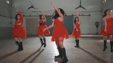 Bir grup kırmızı elbiseli dansçı endüstriyel alanda enerjik bir performans sergiliyorlar. Arka planda bir spot ışığı ve büyük hayranlar var, dinamik bir atmosfer yaratıyorlar..