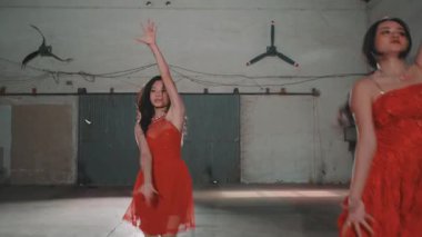 Kırmızı elbiseli iki kadın, sanayi bölgesinde, tavanda hayranlarıyla dans ediyorlar. Arka planda gri bir duvar ve canlı bir atmosfer yaratan bazı ekipmanlar var..