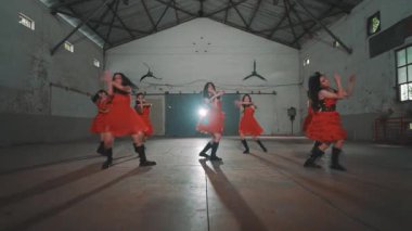 Bir grup kırmızı elbiseli dansçı endüstriyel alanda gösteri yapıyorlar. Sahne onların senkronize hareketlerini ve yerdeki gölgelerini yakalar ve dinamik bir atmosfer yaratır..