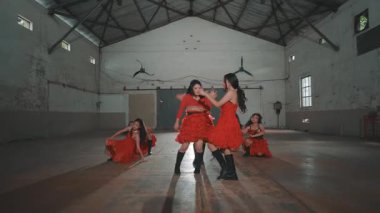 Bir grup kırmızı elbiseli dansçı yüksek tavanlı ve geniş pencereli endüstriyel bir alanda gösteri yapıyorlar. Enerjik koreografi senkronize hareketlerini gösteriyor..