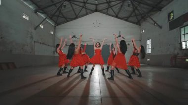 Kırmızı elbiseli altı dansçı yüksek tavanlı ve tahta kirişli endüstriyel bir alanda gösteri yapıyorlar. Ortam minimalist, doğal ışık sahneyi aydınlatıyor..