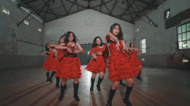 Endüstriyel tarzda geniş bir mekanda dans eden bir grup genç dansçı. Dalgalı ve siyah çizmeli canlı kırmızı elbiseler giyiyorlar, dinamik hareketler sergiliyorlar. Dekor yüksek tavanlar ve çarpıcı ışıklandırmayla korunmasız kirişler içeriyor.