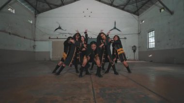 Endüstriyel tarzda geniş bir salonda dans eden bir grup dansçı. Altın aksanlı siyah kıyafetler giyiyorlar, senkronize hareketler sergiliyorlar. Atmosfer enerjik ve dinamik..