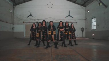 Endüstriyel bir ortamda altı dansçıdan oluşan bir grup. Altın aksanlı siyah kıyafetler giyiyorlar ve güçlü pozlar veriyorlar. Arka planda yüksek tavanlar ve büyük yelpazeler yer alıyor..