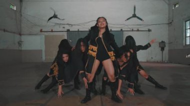 Bir grup siyah kıyafetli ve altın aksanlı dansçı endüstriyel bir alanda dinamik pozlar veriyorlar. Arka planda çıplak bir duvar ve tavan vantilatörü var, dramatik bir atmosfer yaratıyor..