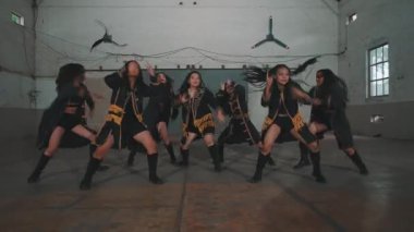 Endüstriyel tarzda geniş bir odada enerjik bir şekilde gösteri yapan bir grup dansçı. Altın aksanlı şık siyah kıyafetler giyiyorlar, senkronize hareketler sergiliyorlar ve neşe ifadeleri sergiliyorlar..