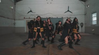 Bir grup dansçı geniş bir endüstriyel ortamda senkronize bir gösteri yapıyorlar. Altın aksanlı siyah kıyafetler giyiyorlar, dinamik hareketler ve ifadeler sergiliyorlar..