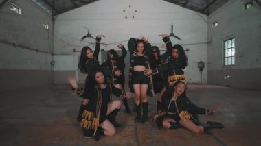 Bir grup dansçı endüstriyel alanda senkronize hareketler yapıyor. Dansçılar siyah kıyafetler giyip sarı aksanlı, dinamik pozlar ve enerji gösteriyorlar. Arka planda yüksek tavanlı geniş, kırsal bir iç mekan var.