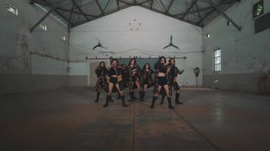 Bir grup dansçı endüstriyel alanda gösteri yapıyor, dinamik hareketler ve koreografi sergiliyorlar. Dansçılar altın aksanlı şık siyah elbiseler giyerek arka planda çarpıcı bir görsel kontrastı oluşturuyorlar..