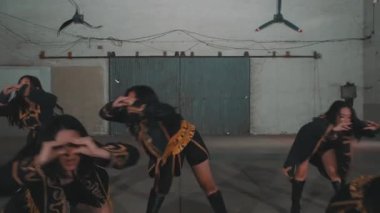 Bir grup siyah kıyafetli ve altın aksanlı dansçı endüstriyel ortamda dramatik bir dans sergiliyorlar. Merkezi dansçı, başkaları tarafından çevrelenmiş anlamlı hareketlerle göze çarpar..