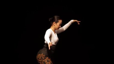 Sahnede zarif bir dansçı, geleneksel bir kıyafetle zarif hareketler sergiliyor. Arka plan koyu, dansçının formunu ve ifadesini vurguluyor..