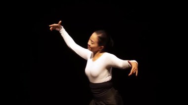 Sahnede zarif bir performans sergileyen bir dansçı karanlık bir arkaplanda karmaşık el hareketleri sergiliyor. Dansçı geleneksel bir kıyafet giyer, kültürel ifade ve sanatın altını çizer..