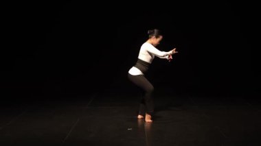 Sahnede siyah bir arka planda sahne alan, etkileyici el hareketleri ve zarif bir duruş sergileyen bir dansçı. Dansçı, akıcılığı ve zarafeti vurgulayan, beyaz ve siyah tayt giyer..
