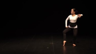 Sahnede sahne ışıkları altında uzun kollu beyaz bir bluz ve siyah tayt giyen bir dansçı. Arka plan karanlık, dansçının hareketlerini ve ifadelerini vurguluyor..