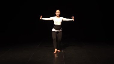 Sahnede dramatik bir duruş sergileyen bir dansçı zarafet ve güç gösterisi sergiliyor. Arka plan karanlık, dansçının hareketlerini ve kıyafetlerini vurguluyor..