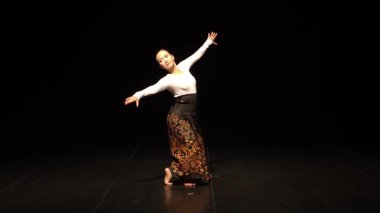 Karanlık bir ortamda sahnede dans eden bir dansçı. Geleneksel, karmaşık desenli bir kıyafet giyiyor. Dansçı zarif bir pozda, akışkan hareketler ve kültürel ifadeler sergiliyor..