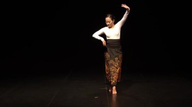 Sahnede dramatik bir şekilde dans eden bir dansçı. Geleneksel ve karmaşık desenli bir kıyafet giyiyor. Arka plan karanlık, dansçının hareketlerini ve ifadesini vurguluyor..