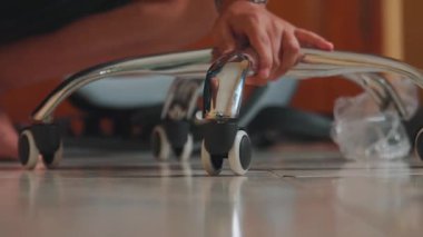 Krom taban ve tekerleklere odaklanarak sandalye monte eden birinin yakın çekimi. Zemin kiremitli ve arka planda sandalyenin diğer parçaları var..