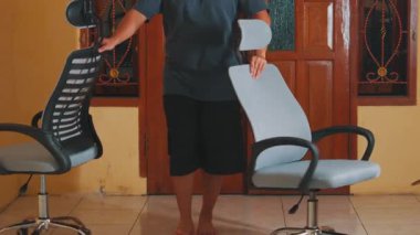 Ahşap kapılı ve fayanslı zemini olan bir odada modern bir ofis sandalyesini ayarlayan biri. Sandalye, ergonomik tasarımı vurgulayan bir ağ sırtı ve ayarlanabilir başlık içerir..