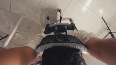 Ön planda modern bir ofis sandalyesini gösteren, tek kolla uzanan bir kişinin yukarıdan aşağıya bakışı. Arka planda üç ayaklı ve düz duvarlı basit bir oda var..