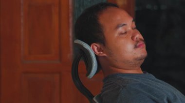 Sandalyede oturan bir adam, elini yüzünü korumak için kaldırmış, muhtemelen bir anlık bir düşünce ya da hayal kırıklığı içinde. Arkaplanda ahşap paneller var, bu da evin içinde rahat bir ortam olduğunu gösteriyor..