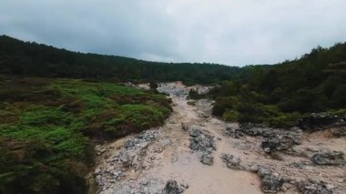 Kuru bir nehir yatağı olan kayalık bir arazinin panoramik görüntüsü, yemyeşil bitki örtüsü ve bulutlu bir gökyüzünün altındaki ağaçlarla çevrili..