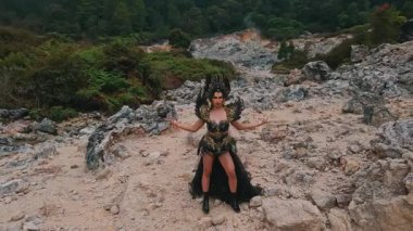 Çarpıcı kostümlü dramatik bir figür kayalık ve çorak bir arazide duruyor. Kıyafet, karmaşık tasarımlar ve doğal çevreyle çelişen cesur bir başlık içeriyor..